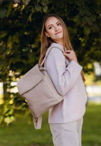 Světlý béžový kožený batoh s klopou, nastavitelnými popruhy a decentně embosovaným logem. Nesený přes pastelově růžový svetr, obklopený zelení.