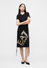 Μαύρο βαμβακερό φόρεμα με κοντά μανίκια, διακοσμημένο με διαγώνιο χρυσό τύπωμα που γράφει "EAT EMPORIO ARMANI" και κορδόνια στο τελείωμα.