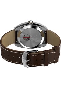 Timex ur med et hus i rustfrit stål, brun læderrem, cremefarvet syning og en gennemsigtig bagside, der viser mekanismen.
