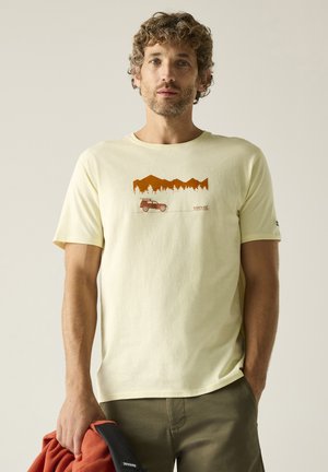 Hombre con cabello rizado que lleva una camiseta amarillo claro con un estampado de montaña y coche, sosteniendo una chaqueta naranja y negra en la mano izquierda.