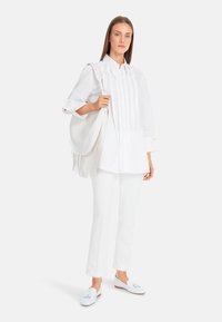 White long-sleeve shirt with pleated front, paired with white trousers and light blue loafers. The model holds a white bag with fringe detail.