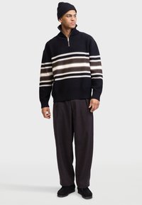 Mann trägt eine schwarze Mütze, schwarzen Reißverschluss-Pullover mit weißen und braunen Streifen, lockere dunkle Hose und schwarze Schuhe, steht vor weißem Hintergrund.