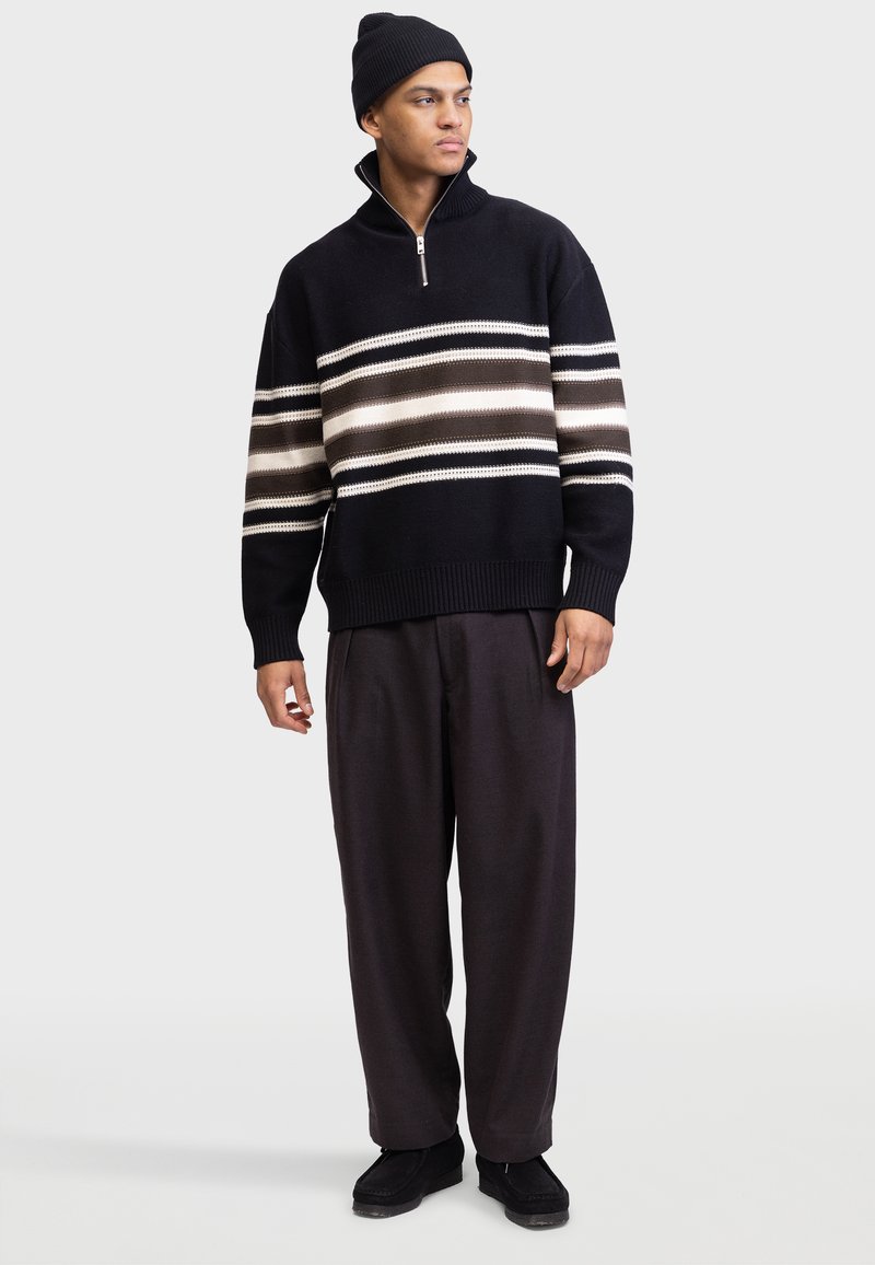 Mann trägt eine schwarze Mütze, schwarzen Reißverschluss-Pullover mit weißen und braunen Streifen, lockere dunkle Hose und schwarze Schuhe, steht vor weißem Hintergrund.