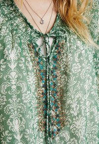 Grönmönstrad blus med knytning vid halsen, med intrikata blå och guld pärlaccent på bröstet, samt en strukturerad tyg.