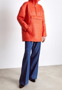 Osoba ubrana w pomarańczową pikowaną kurtkę z kapturem i kieszenią z przodu oraz niebieskie szerokie spodnie jeansowe, stojąca na jasnoszarej podłodze na tle białej ściany.