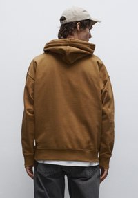 Felpa marrone con vestibilità comoda, grande cappuccio, polsini e orlo a costine, indossata sopra una maglietta bianca, abbinata a jeans grigi e un cappello beige.