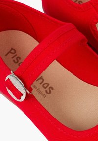 Zapatilla de lona roja con una correa de tela y detalle de hebilla plateada. El interior presenta una plantilla suave de color natural con el texto de la marca.