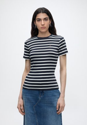 Žena s dlhými tmavými vlasmi, nosiace strieborné kruhové náušnice, námornícky modré a bielo pruhované tričko s krátkym rukávom a modrú rifľovú sukňu, stojaca pred bielym pozadím.