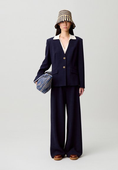 Abito in lana blu navy con camicia bianca con colletto, pantaloni a gamba larga e chiusura con bottoni. Abbinato a un cappello a secchiello a righe e una borsa a tracolla con motivo pattern.