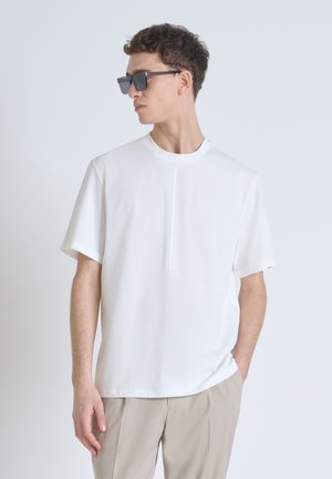 Camiseta blanca de manga corta en tejido ligero con un diseño minimalista y un detalle de costura vertical en la parte delantera. Se lleva con gafas de sol grises.