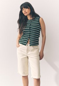 Haut sans manches vert rayé avec boutons à l'avant, associé à un short beige clair retroussé, montrant une tenue décontractée d'été.