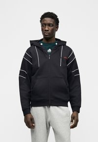 Άνδρας που φοράει μαύρο φούτερ με φερμουάρ και λευκή περιέλιξη, σκούρο πράσινο πουκάμισο Adidas και ανοιχτό γκρι φόρμα, σε απλό λευκό φόντο.