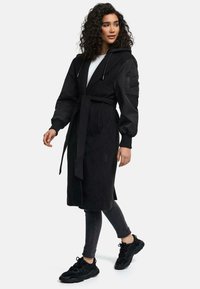 Black long coat with a hood, cinched waist, and patch pockets. Features contrasting fabric on sleeves and a relaxed fit. Paired with black sneakers.