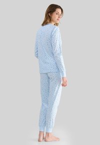 Mujer de pie descalza, vestida con una blusa de manga larga azul clara y pantalones con un pequeño patrón floral azul oscuro sobre un fondo gris.