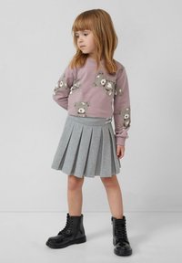 Junges Mädchen mit roten Haaren, das einen lila Teddybär-Sweatshirt, einen grauen Faltenrock und schwarze Schnürstiefel trägt, steht vor einem schlichten Hintergrund.