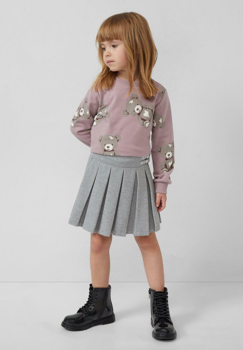 Junges Mädchen mit roten Haaren, das einen lila Teddybär-Sweatshirt, einen grauen Faltenrock und schwarze Schnürstiefel trägt, steht vor einem schlichten Hintergrund.