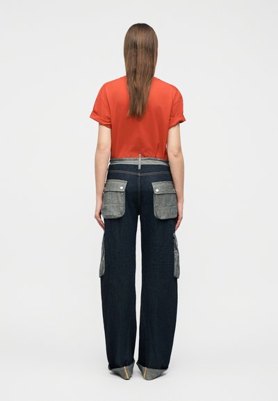 Άτομο με μακριά καστανά μαλλιά, που φοράει κόκκινο κοντομάνικο πουκάμισο και σκούρο παντελόνι με φαρδιά πόδια και μεγάλες γκρι τσέπες με παρόμοια καλύμματα, όρθιο με την πλάτη προς τα εμάς.