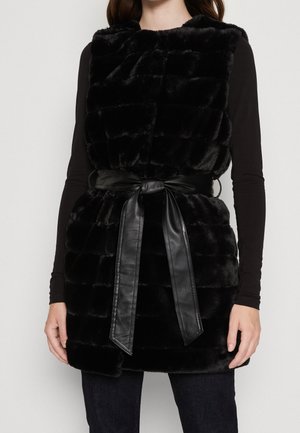 Femme portant un gilet sans manches en fausse fourrure noire avec une large ceinture en cuir noire nouée en nœud à la taille.