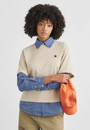 Donna con capelli ricci che indossa una t-shirt beige a strati sopra una camicia a righe blu, pantaloni kaki, che tiene un berretto da baseball arancione in una mano.