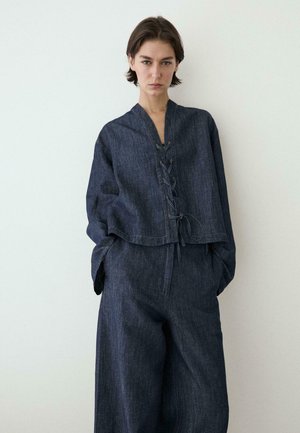 Femme debout contre un mur blanc, vêtue d'un haut en denim bleu foncé, ample, avec un laçage à l'avant, et d'un pantalon ample assorti. Mains dans les poches.