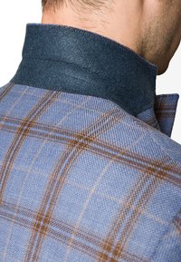 Niebieska tkanina w kratę z brązowymi i niebieskimi wzorami. Ciemny aksamitny kołnierz z widocznym szwem. Zbliżenie, które koncentruje się na fakturze i wzornictwie.