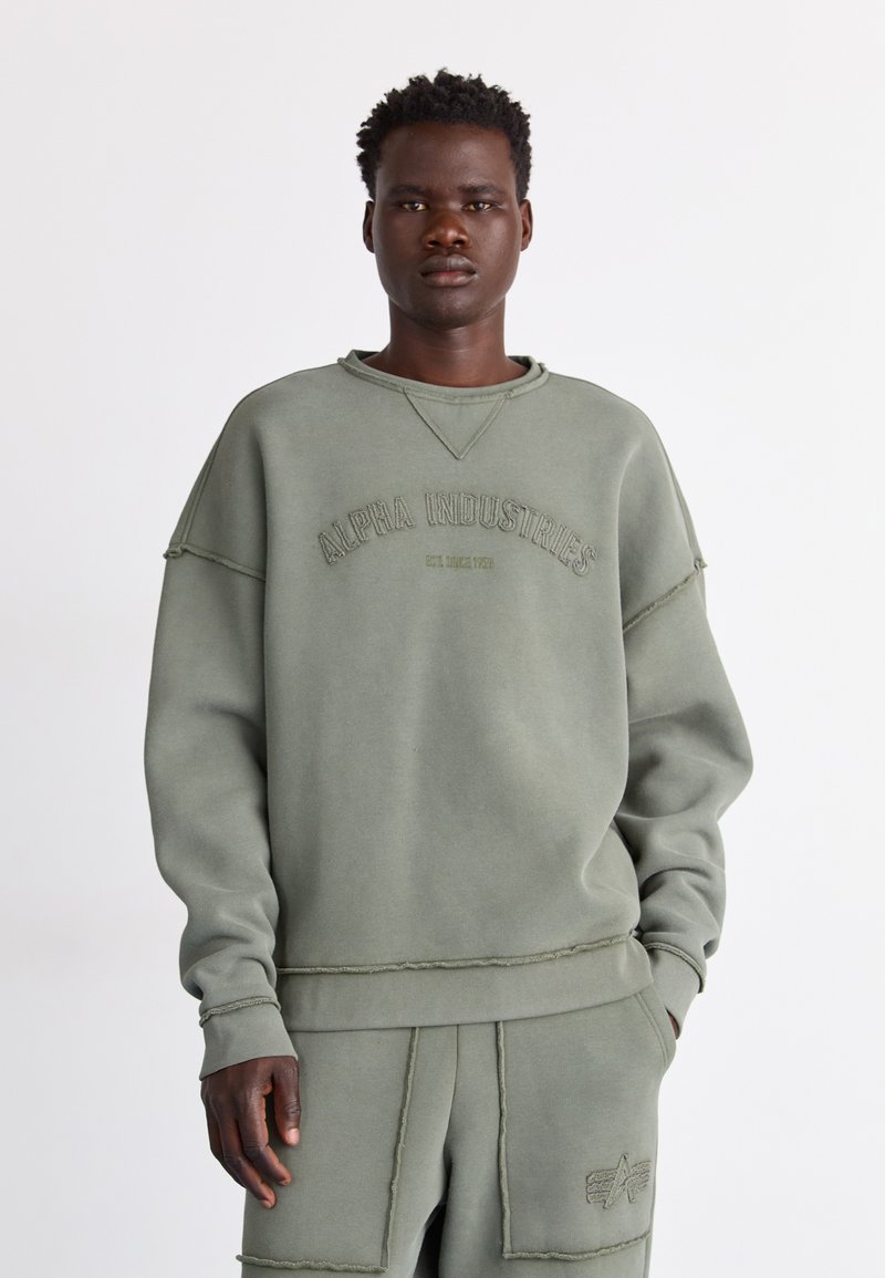 Olivenfarget oversized hettegenser med ribbestrikket hals og råskåret detaljer; har brodert "Alpha Industries" logo på forsiden.