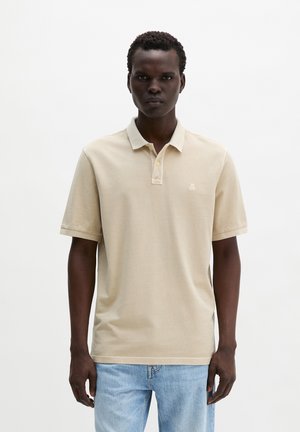 Man staat en draagt een beige poloshirt met korte mouwen en lichtblauwe spijkerbroek tegen een effen witte achtergrond.