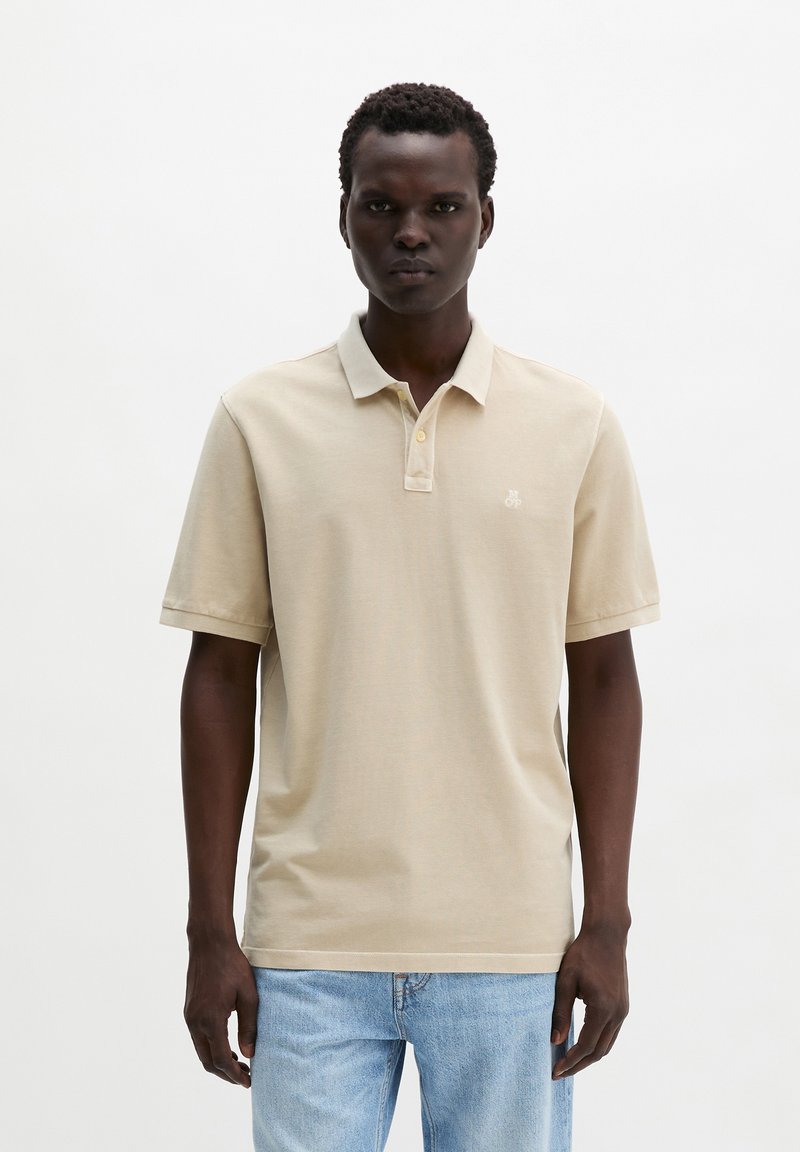 Homme debout portant un polo à manches courtes beige et un jean bleu clair, sur fond blanc uni.