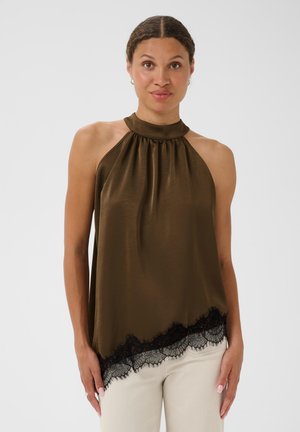 Femme portant un top sans manches vert olive à col halter avec une bordure en dentelle noire à l'ourlet et un pantalon de couleur claire sur un fond uni.