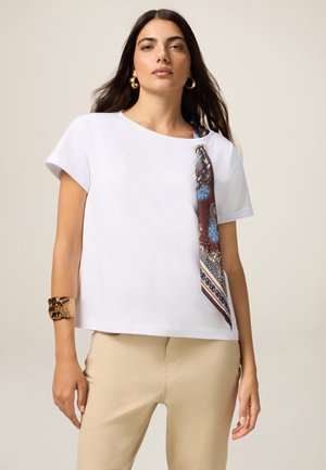 Witte katoenen T-shirt met korte mouwen, met een decoratieve bruin en blauwe geborduurde sjaal die aan de linkerschouder is geknoopt. Beige broek zichtbaar.