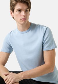 Light blue cotton t-shirt with short sleeves, round neckline, and a smooth texture, worn by a seated person against a plain background.