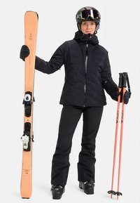 Oseba v črnem smučarskem kompletu, s čelado in očali, drži breskevovo smučanje v levi roki in ustrezne smučarske palice v desni roki.