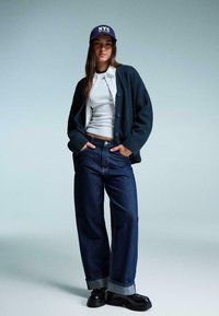 Navy svetr, bílý tričko, volné tmavé džínové kalhoty s ohrnutými nohavicemi, černé boty a námořnická čepice s textem "NYC".