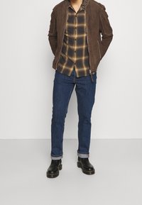 Chaqueta marrón de ante con cremallera sobre una camisa de cuadros marrón y amarilla, combinada con vaqueros azul oscuro y botas de cuero negro.