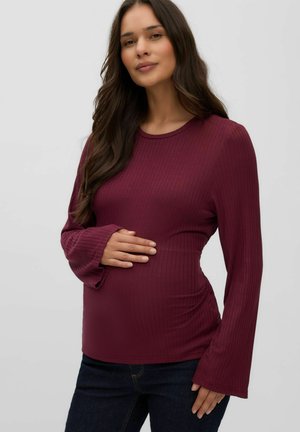 Femme enceinte aux longs cheveux foncés, portant un haut bordeaux côtelé à manches longues et un jean foncé, tenant doucement son ventre et regardant l'appareil photo.