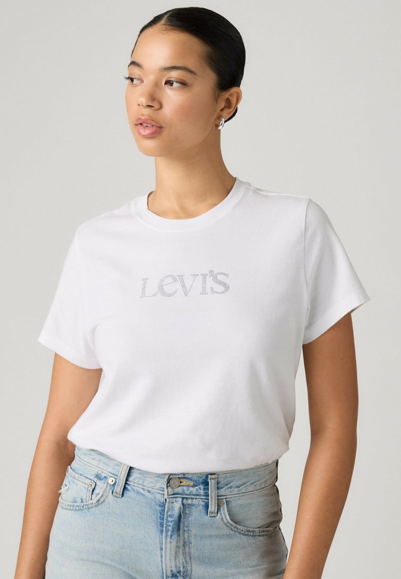 Jeune femme aux cheveux foncés portant un t-shirt blanc Levi's et un jean bleu clair, regardant sur le côté devant un fond clair uni.