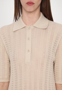 Femme portant un polo en maille beige à manches courtes avec des rayures texturées verticales et trois boutons sur la patte de boutonnage.