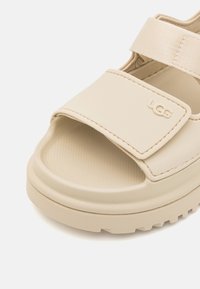 Bež UGG suss, mille rihmaga, tekstureeritud tallaga ja reguleeritava Velcro kinnitusega. Lame disain ümarate varvastega ja paksu tallaga.