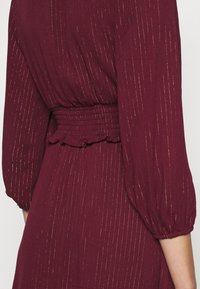Robe bordeaux en tissu texturé, avec des rayures verticales dorées, une taille froncée et des manches longues bouffantes élastiquées.
