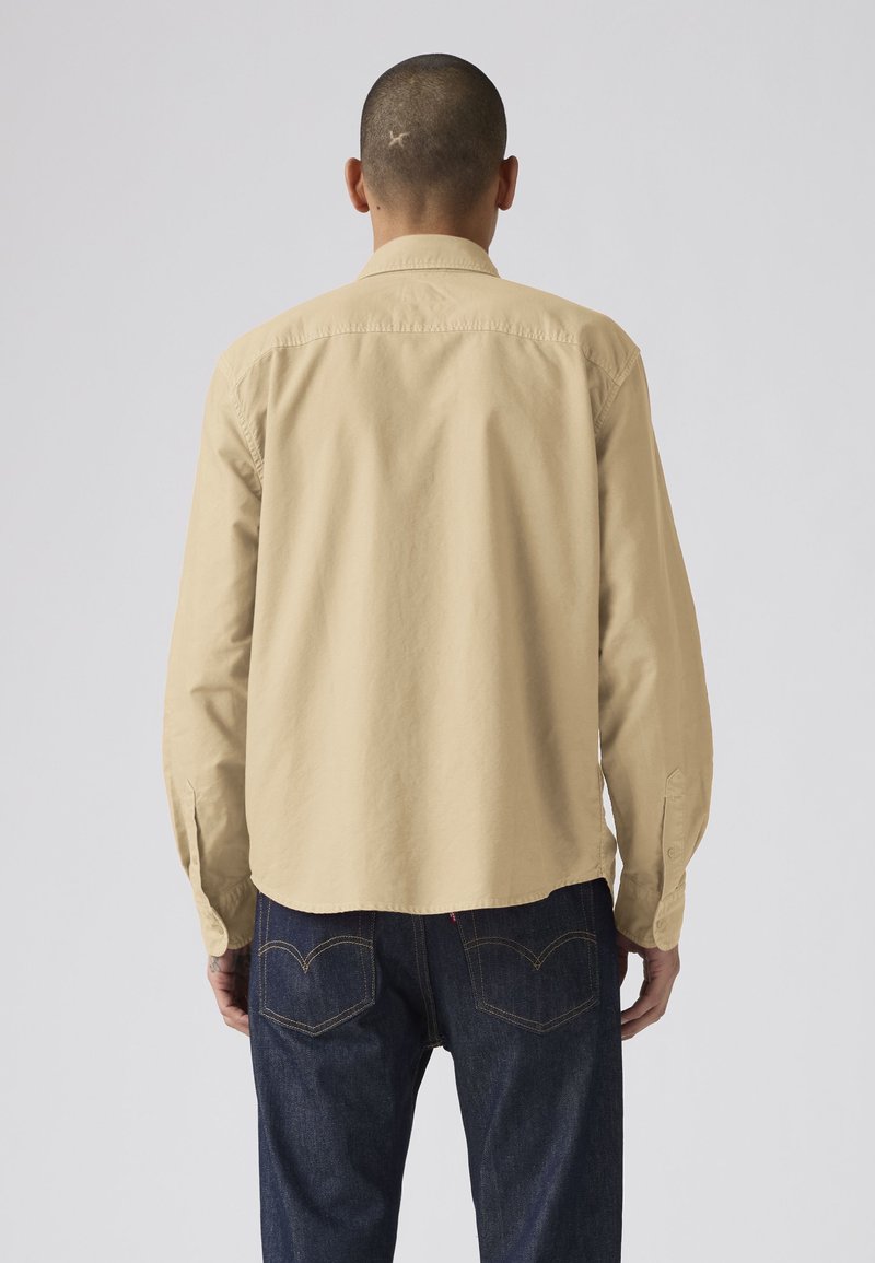Mann mit rasiertem Kopf trägt ein beigefarbenes langärmeliges Hemd und dunkelblaue Jeans und steht mit dem Rücken zu einem einfarbigen hellgrauen Hintergrund.