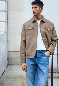 Brun knap-tryk jakke med to brystlommer, lavet af tekstureret stof; parret med lyseblå denim jeans og en hvid skjorte.