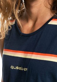 Gros plan sur une personne portant un haut sans manches bleu marine avec des rayures horizontales rouges, orange et crème, et le logo "Quiksilver" sur la poitrine.