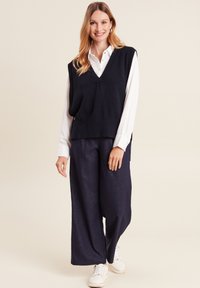 Marineblauw gebreid vest met een V-hals, gelaagd over een wit shirt, gecombineerd met donkere fijngestreepte wijde broek en witte sneakers.