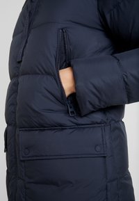 Chaqueta de plumón azul marino con bolsillo con cremallera, textura suave, diseño acolchado y puños ajustables. Presenta un bolsillo con solapa abotonado en la parte frontal.