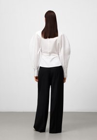 Witte katoenen blouse met oversized pofmouwen, getailleerde taille en knoopmanchetten, gecombineerd met zwarte wijde broekspijpen. Effen achtergrond.