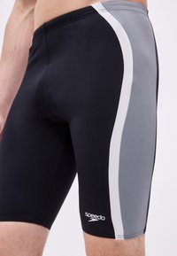 Άνδρας που φοράει μαύρα και γκρι μαγιό Speedo τύπου jammers με λευκή ρίγα στο πλάι, εμφανιζόμενος από τη μέση έως το μέσο του μηρού σε απλό φόντο.
