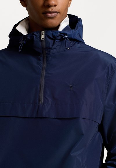 Veste imperméable bleu marine avec une demi-fermeture éclair, un col montant et des cordons de serrage ajustables, arborant un petit logo sur la poitrine.