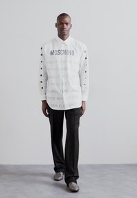 Cămașă albă cu text imprimat și buline negre pe mâneci, asociată cu pantaloni negri și pantofi cu un model. Material neted, croială ajustată.