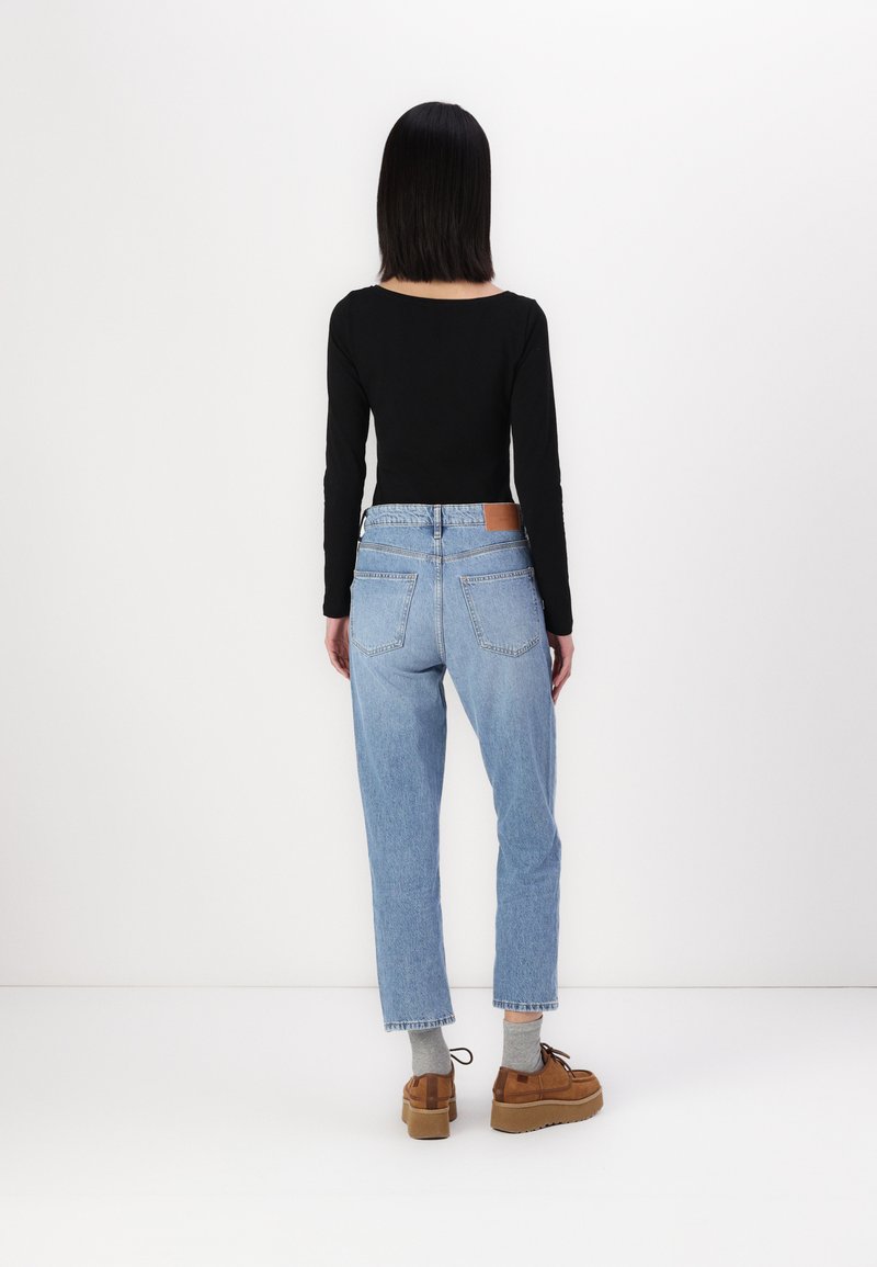 Schwarzes langärmliges Oberteil kombiniert mit hellblauen hochgeschnittenen Jeans, die auf der Rückseite ein braunes Lederlabel tragen, sowie braunen Plateau-Schuhen.