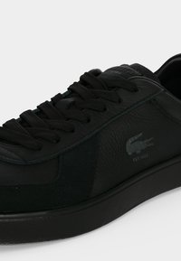 Gros plan sur une basket en cuir noir avec des lacets noirs, montrant des panneaux latéraux texturés et un petit logo crocodile avec le texte "EST 1933".